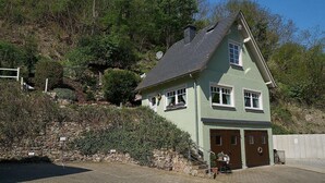 A two-story house with a steeply pitched roof, a stone retaining wall, and a paved driveway leading to a garage.