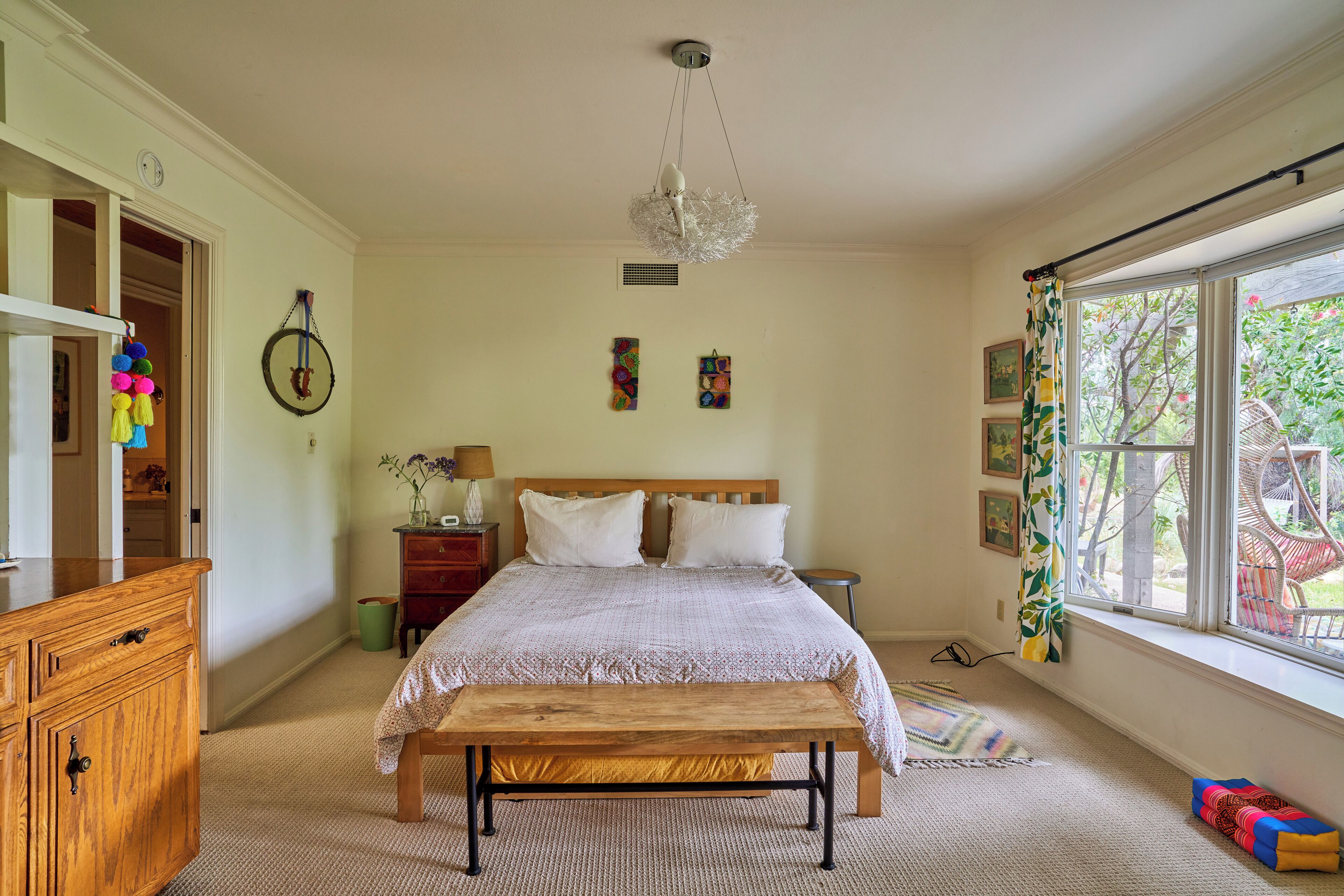 A bedroom with a wooden bed, a bench, a dresser, a window with a view, and a hanging light fixture.