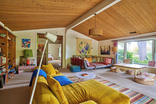 Un salón acogedor con un sofá amarillo, techo de madera y un ventanal con vistas a la vegetación.