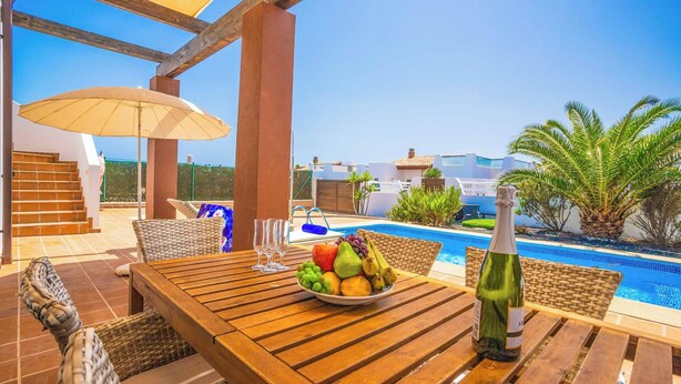 Un área de comedor al aire libre con una mesa de madera, sillas de mimbre, un cuenco de frutas, una botella de champán y una piscina al fondo.