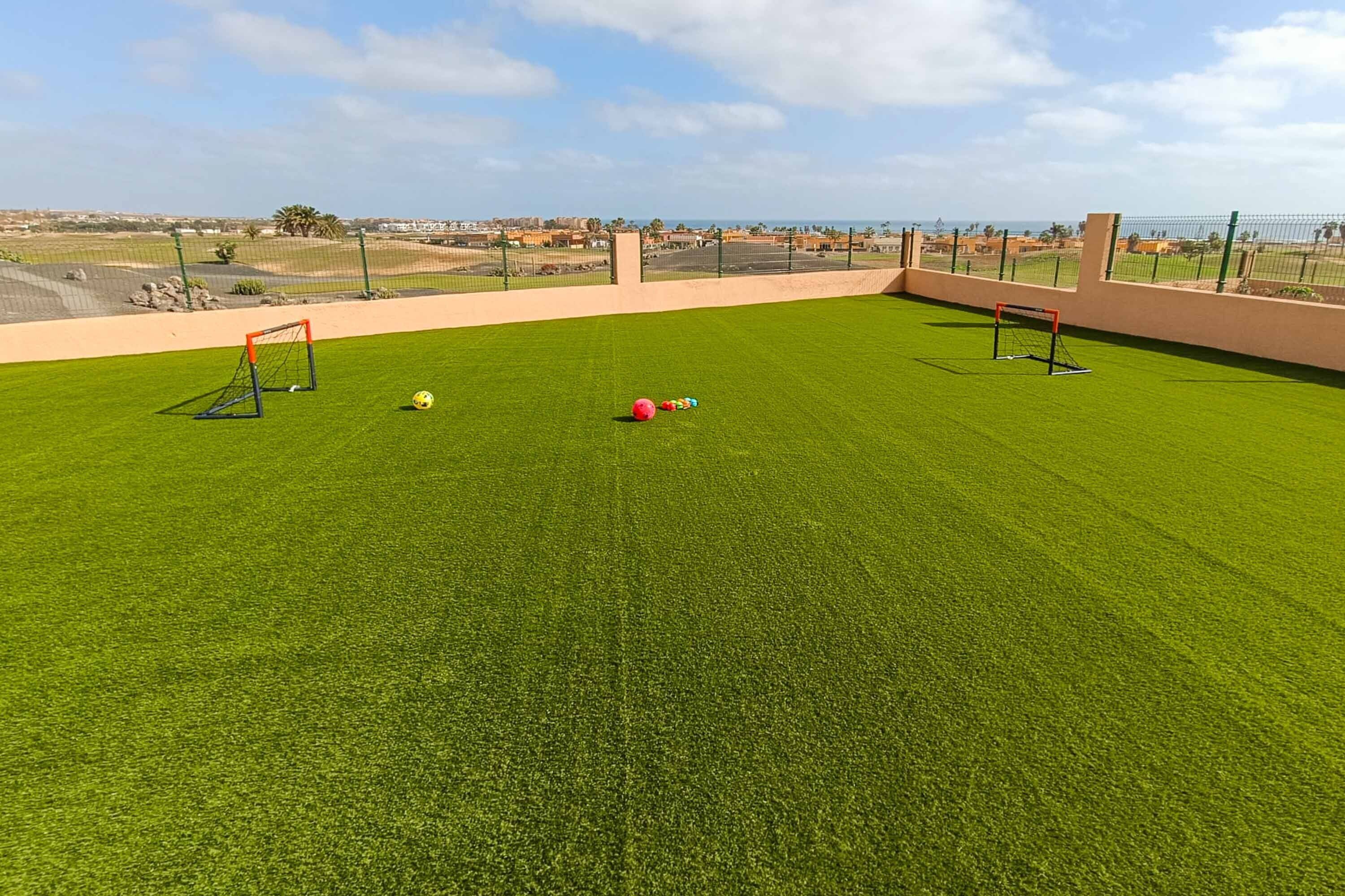 A well-maintained artificial grass field with two soccer goals and a net, surrounded by a fence, with a clear view of the distant landscape.