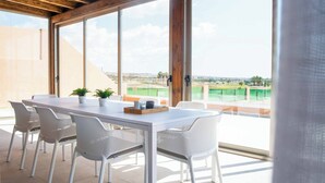 Un coin repas avec une table et des chaises blanches, de grandes fenêtres et une vue sur un court de tennis.