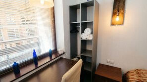 Une chambre d’hôtel dotée d’une grande fenêtre avec stores, d’une table en bois sur laquelle sont posées deux bouteilles bleues, d’une étagère grise où sont rangées des serviettes et d’un luminaire fixé au mur.