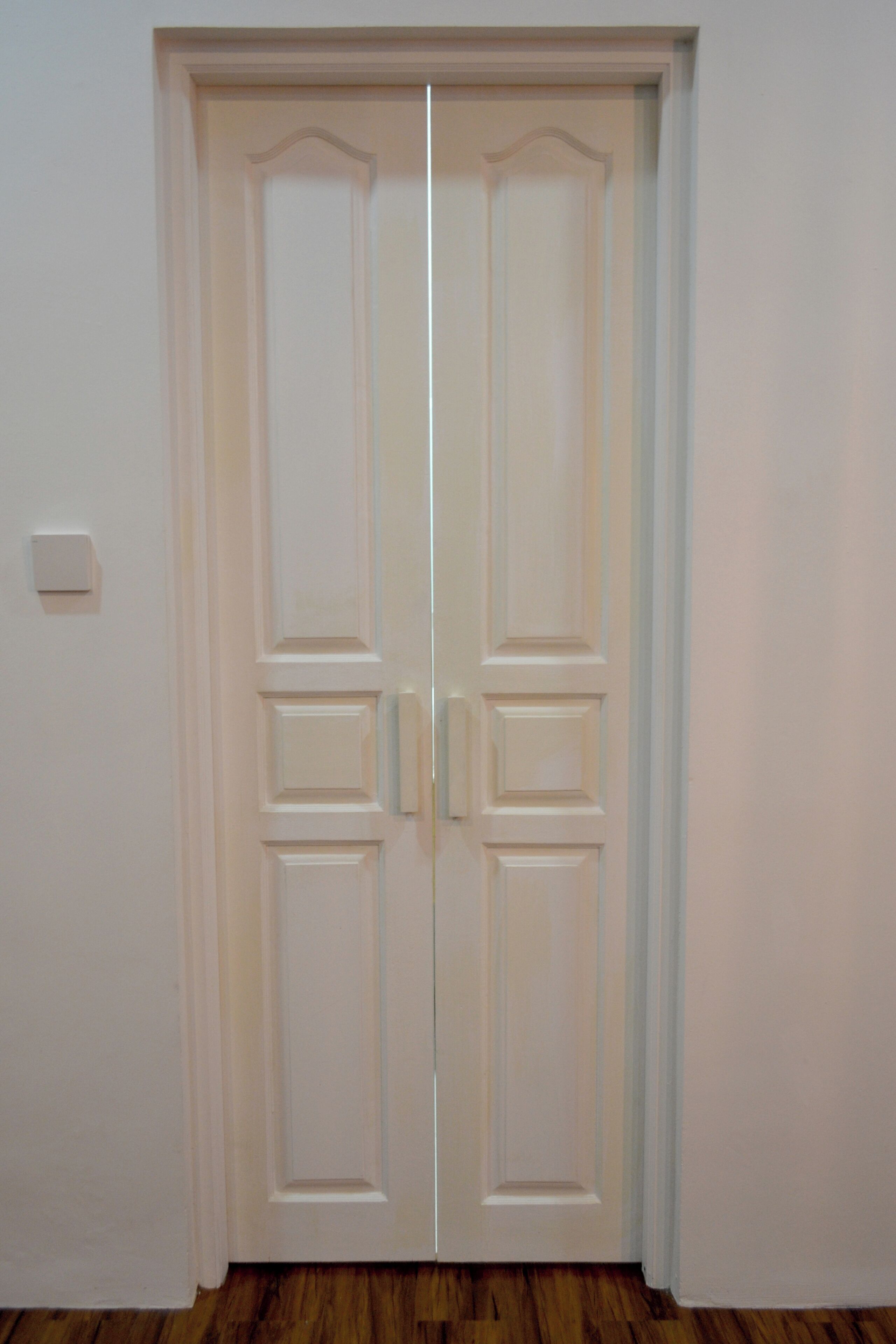 Puerta doble blanca con un interruptor de luz en la pared a la izquierda.