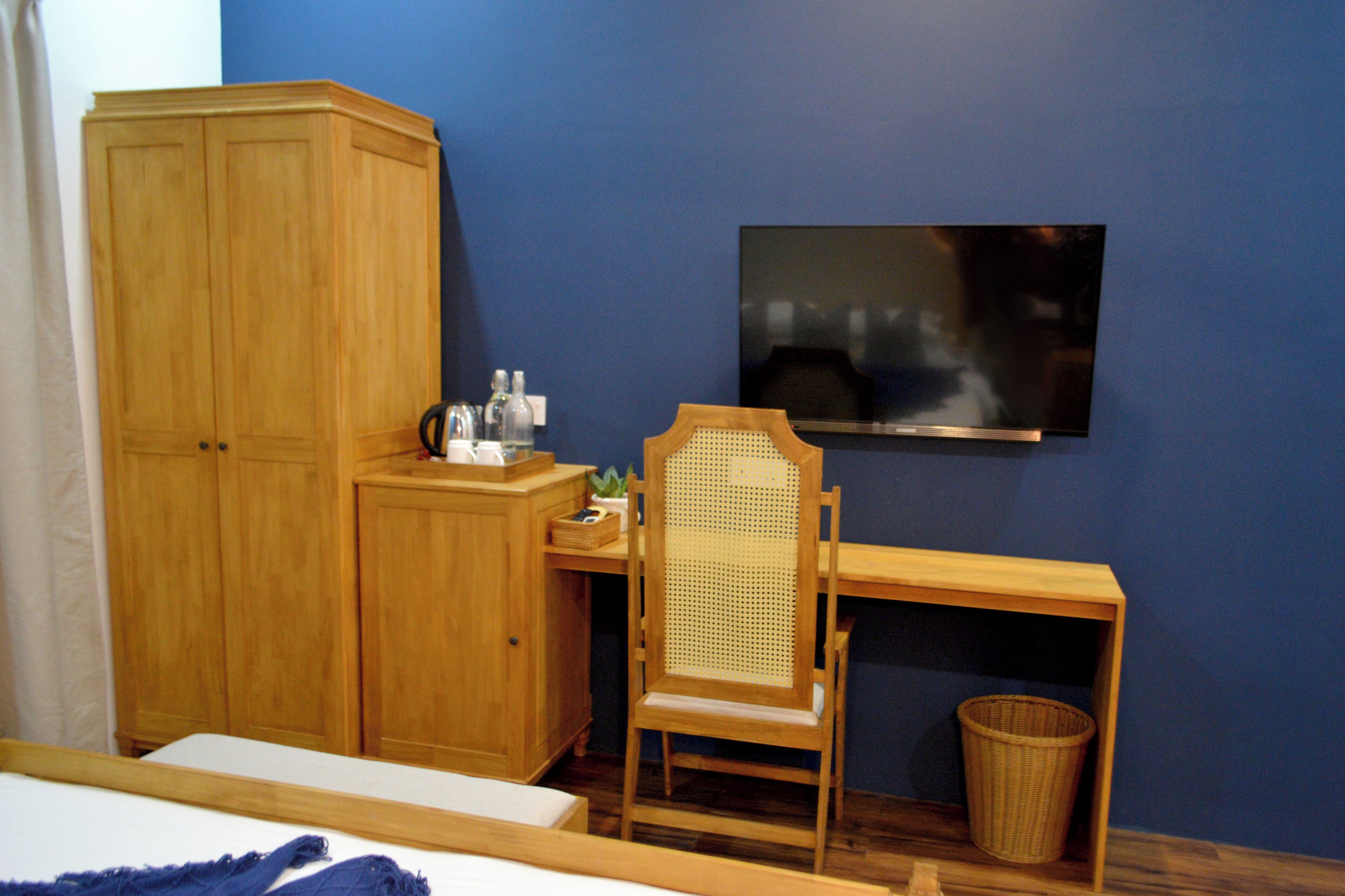 Un dormitorio con un armario de madera, un escritorio con silla, un televisor en la pared y una pequeña cesta de mimbre.