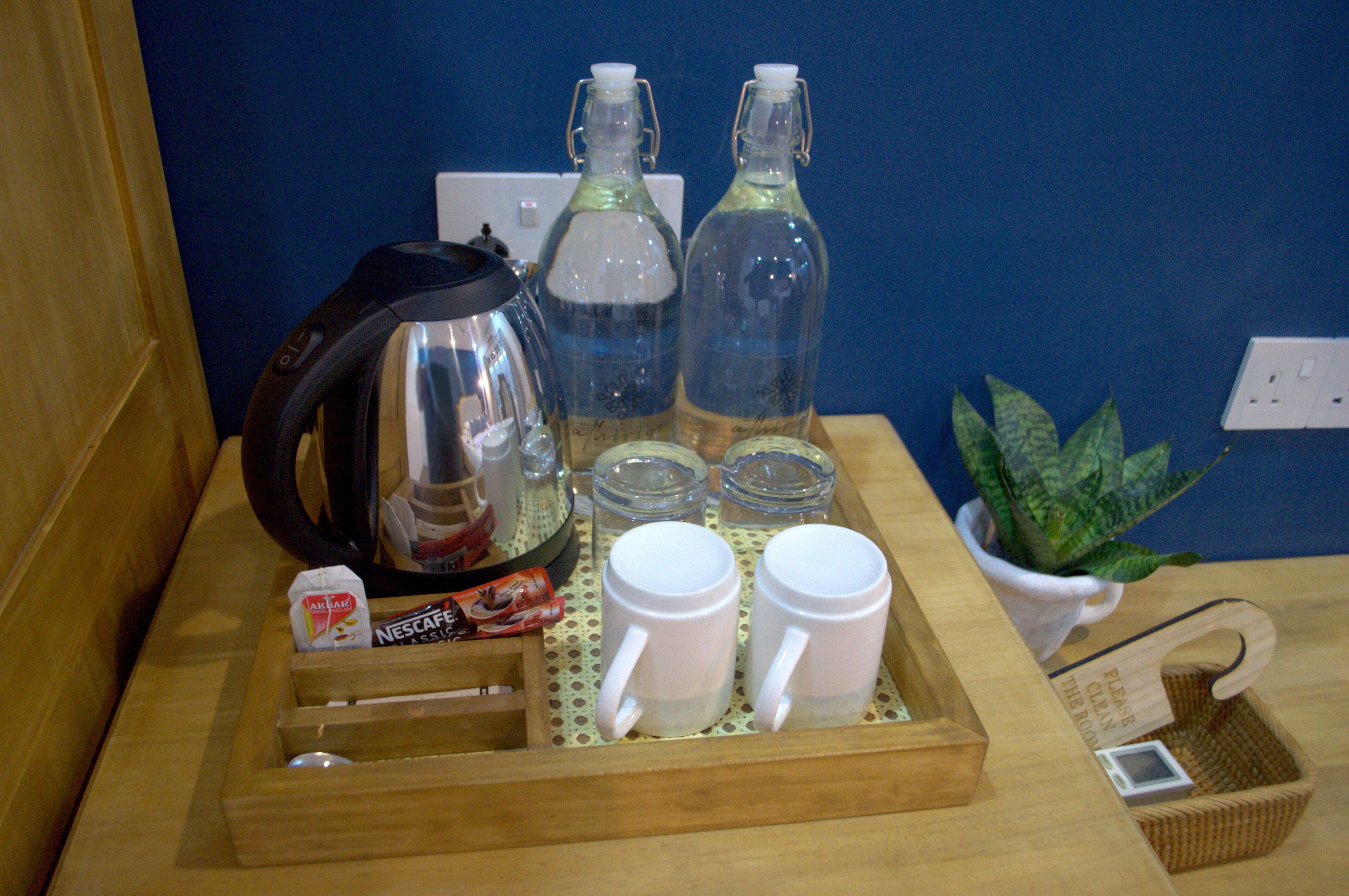 Un plateau en bois avec une bouilloire, deux bouteilles, deux tasses, une plante et un panier sur lequel est posée une pancarte.