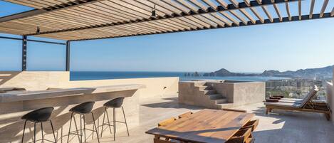 Una terraza en la azotea con una pérgola de madera, una mesa de madera con sillas y vistas al mar y al paisaje urbano.