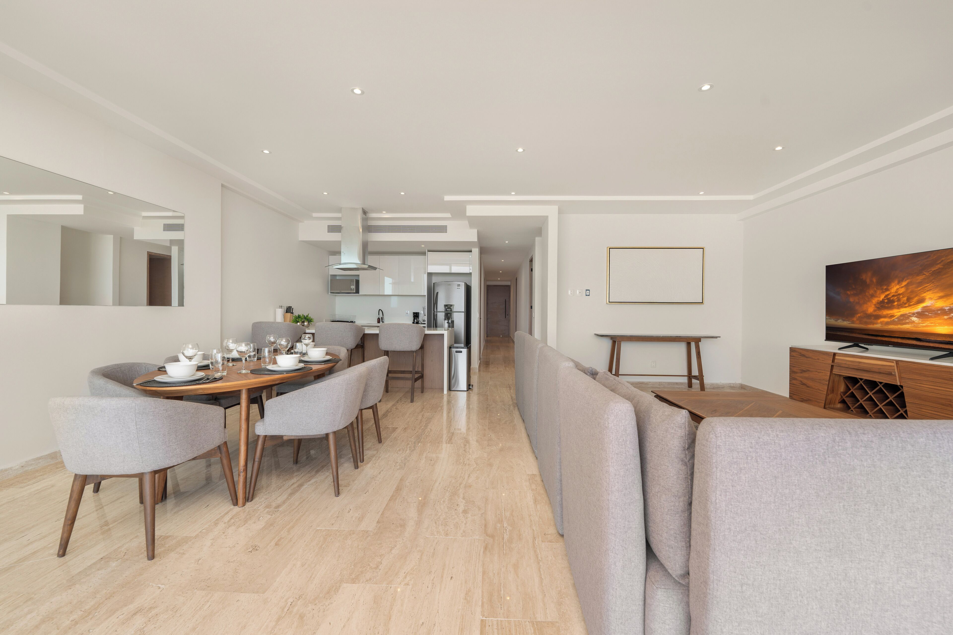 Un espace repas moderne avec une table en bois et des chaises à revêtement gris, une cuisine en arrière-plan et un téléviseur posé sur un meuble en bois.
