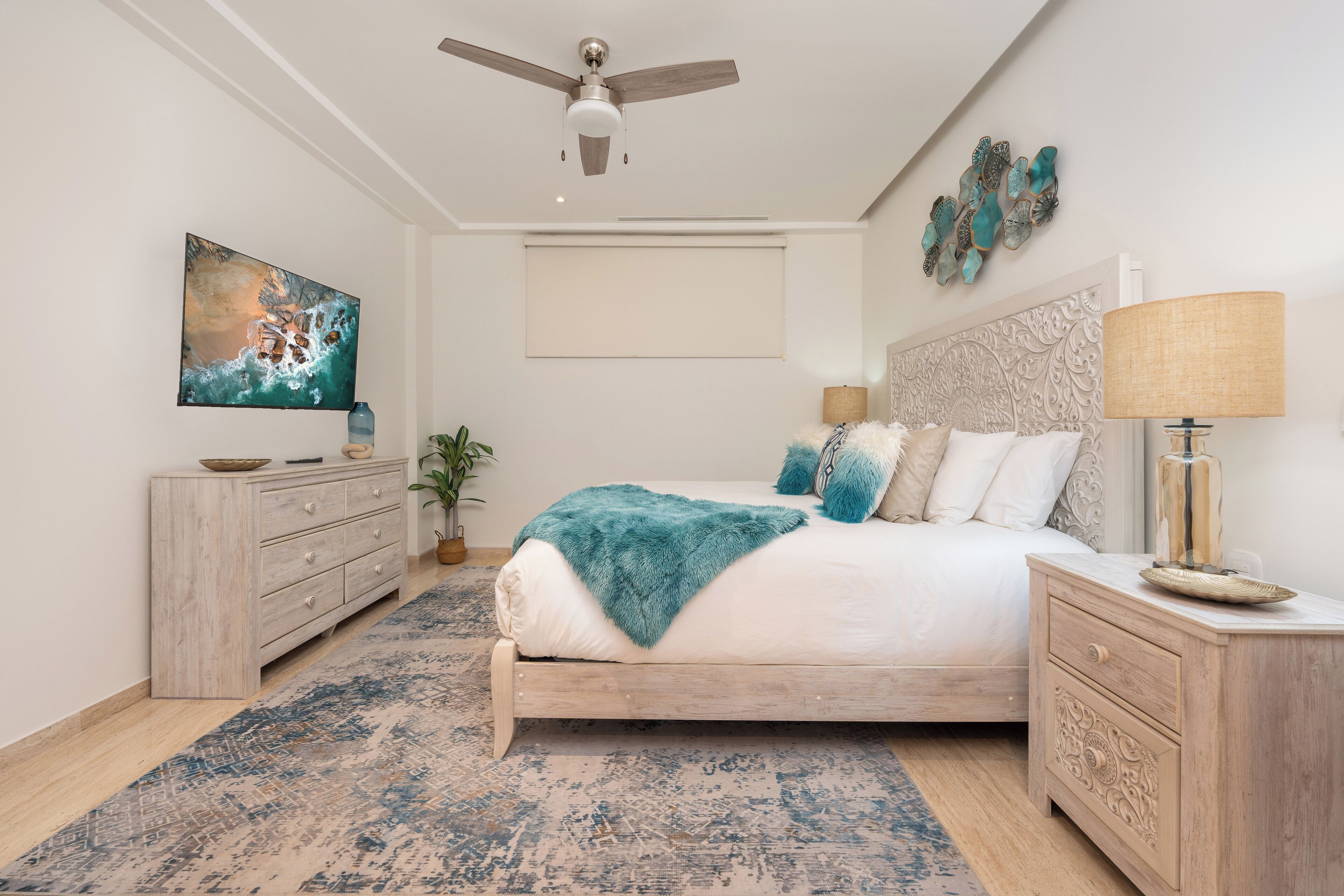 Un dormitorio con cama, ventilador de techo, televisor, un adorno en la pared, mesita de noche y mesilla.