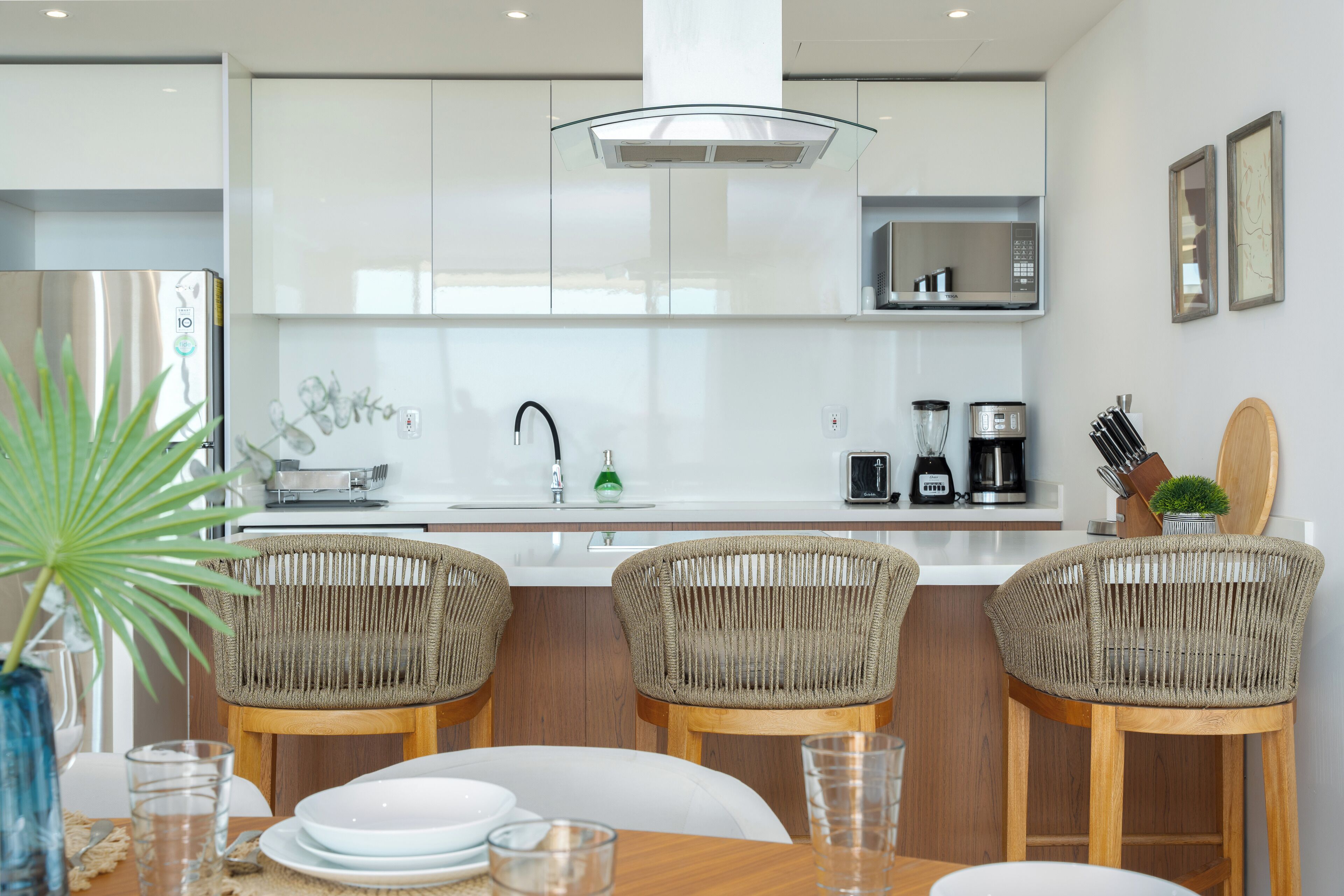 A modern kitchen with white cabinets, a stainless steel refrigerator, a built-in oven, and a coffee maker. There are wicker chairs and a wooden table with plates and glasses.