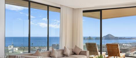 Una habitación de hotel moderna con un balcón amplio, un sofá, una mesa de comedor y vistas al mar y al paisaje urbano.
