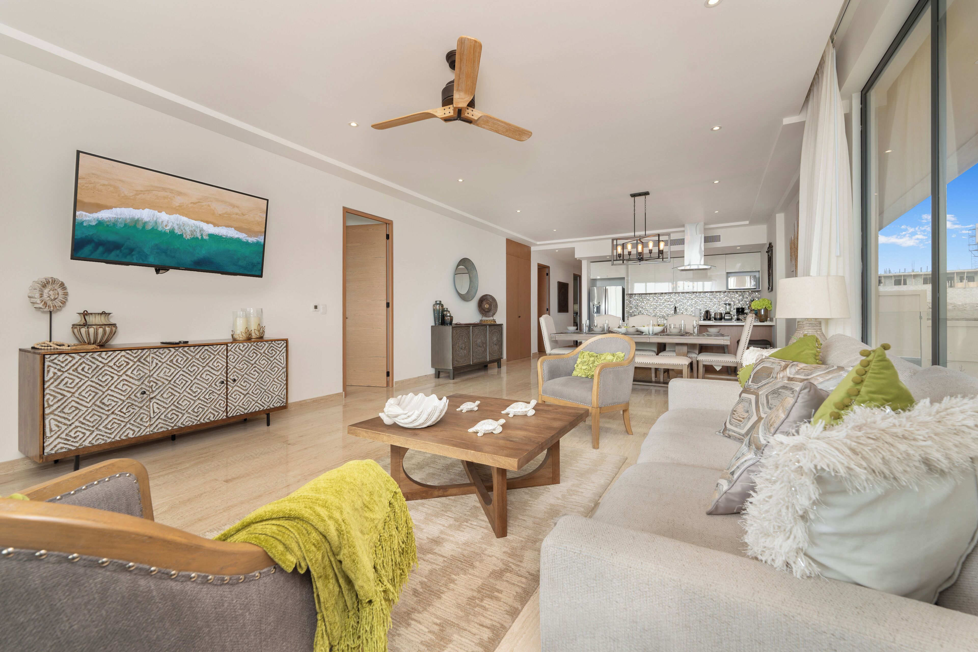 A modern living room with a flat-screen TV, a wooden ceiling fan, and a dining area with a kitchen in the background.