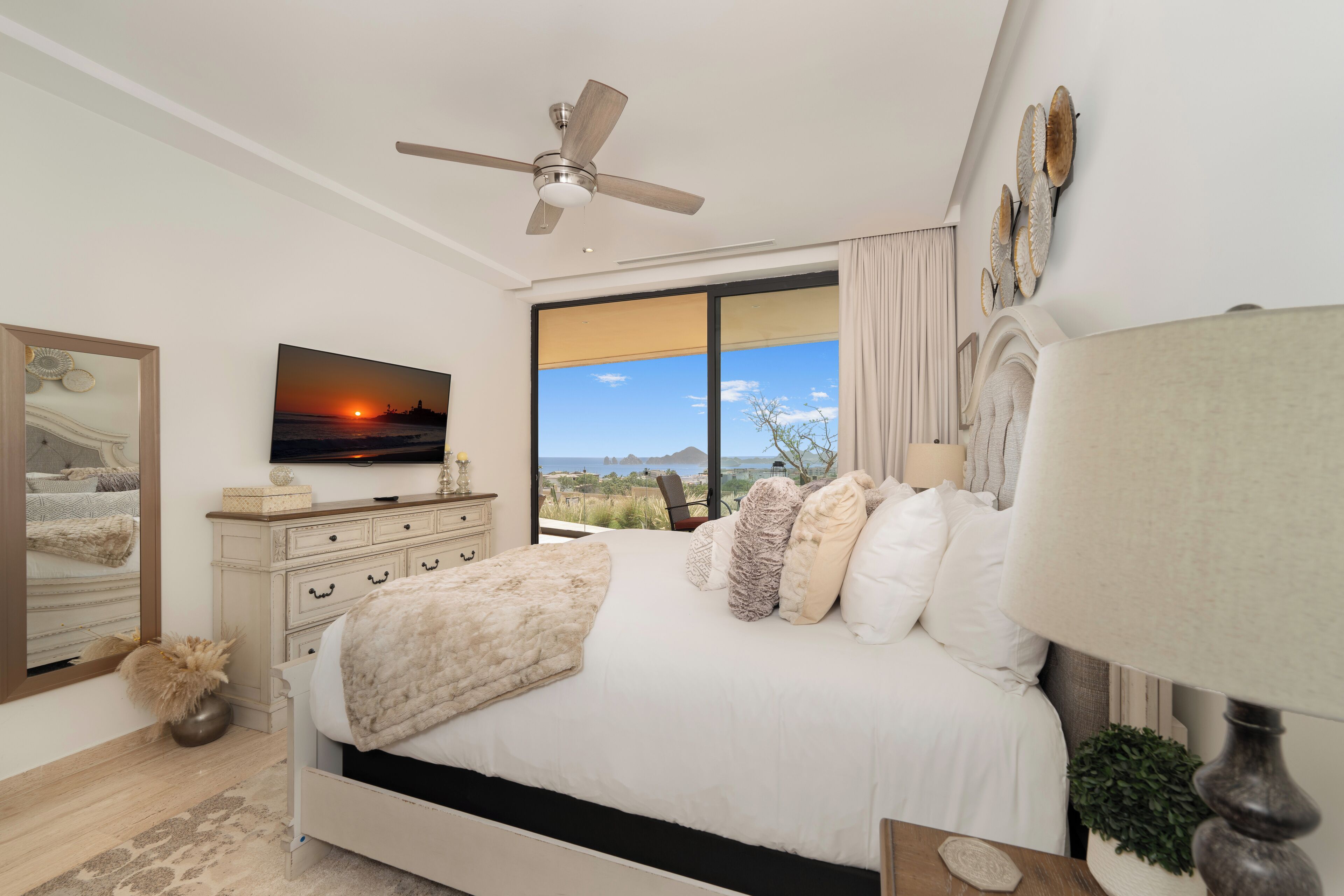A bedroom with a bed, a ceiling fan, a TV, a large window with a view, and a dresser.