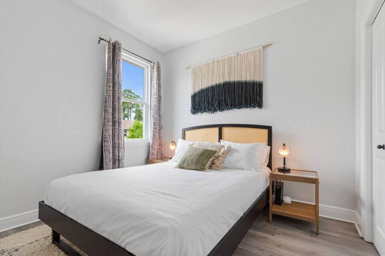 A bedroom with a large bed, a bedside table with a lamp, a window with curtains, and a wall-mounted tapestry.