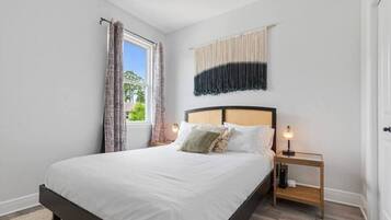 A bedroom with a large bed, a bedside table with a lamp, a window with curtains, and a wall-mounted tapestry.