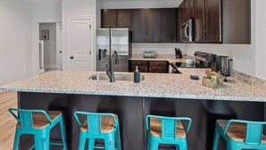 Una cocina moderna con armarios de madera oscura, electrodomésticos de acero inoxidable, encimera de granito y tres taburetes de bar color turquesa.