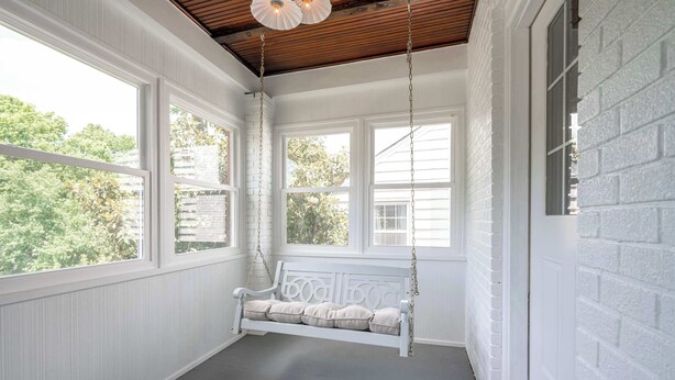 Una habitación con paredes blancas y techo de madera, un banco mecedor blanco con cojines, y ventanas amplias que dan a vistas de árboles.
