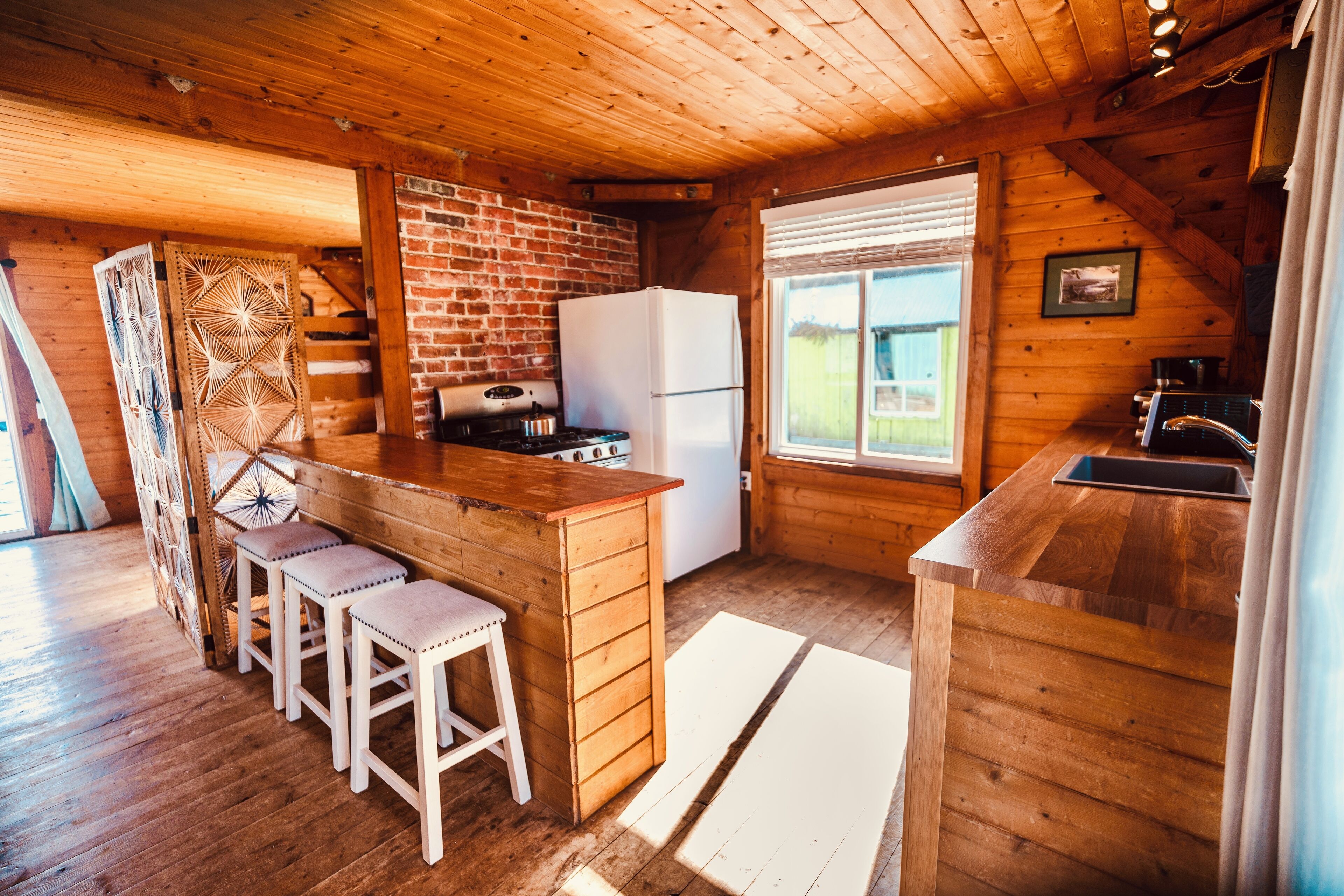 A wooden cabin kitchen with a brick wall, wooden bar stools, a refrigerator, and a window with blinds.