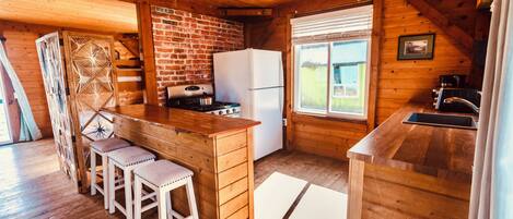 A wooden cabin kitchen with a brick wall, wooden bar stools, a refrigerator, and a window with blinds.