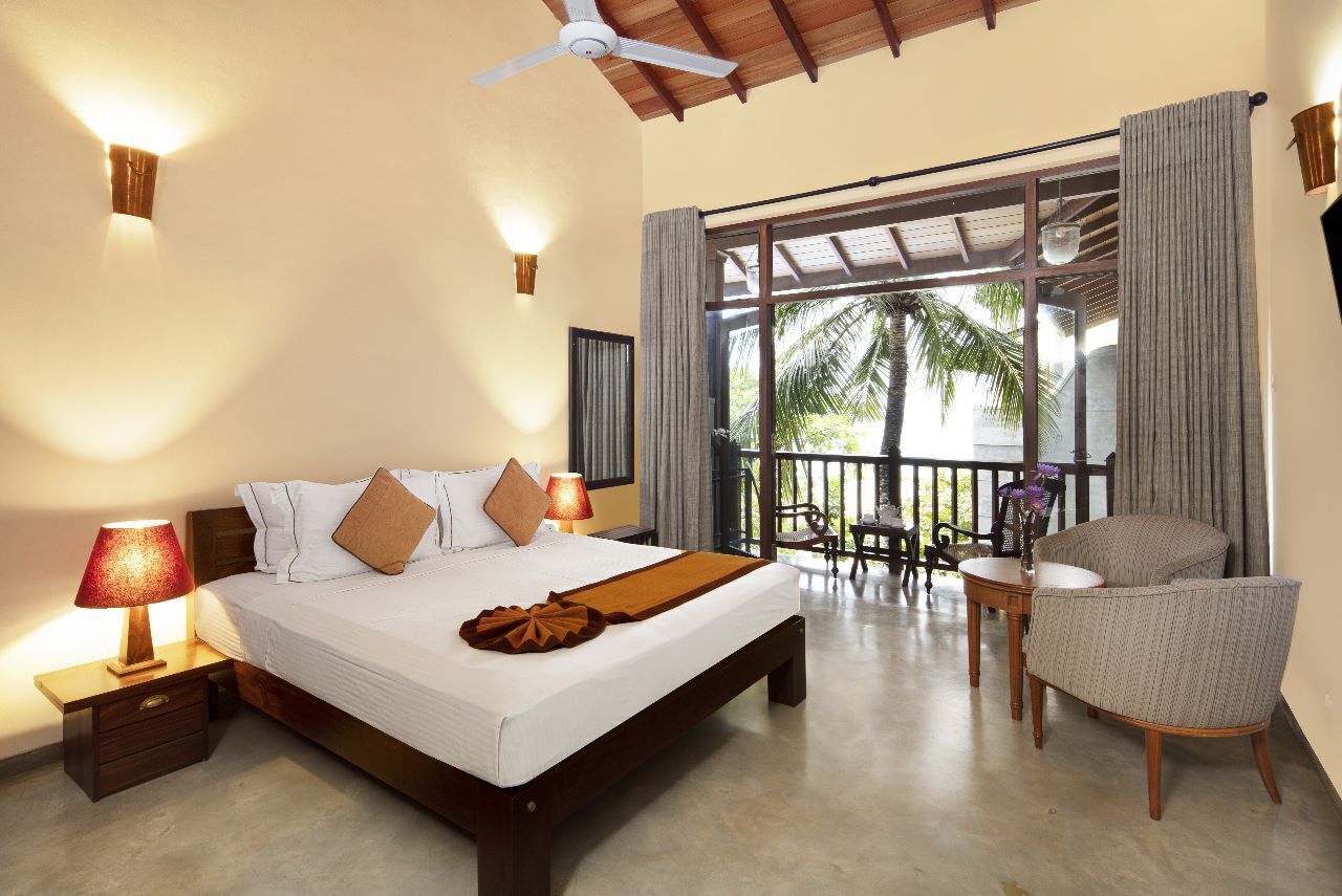 Een hotelkamer met een groot bed, een plafondventilator, een balkon met een tafel en stoelen, en uitzicht op palmbomen.