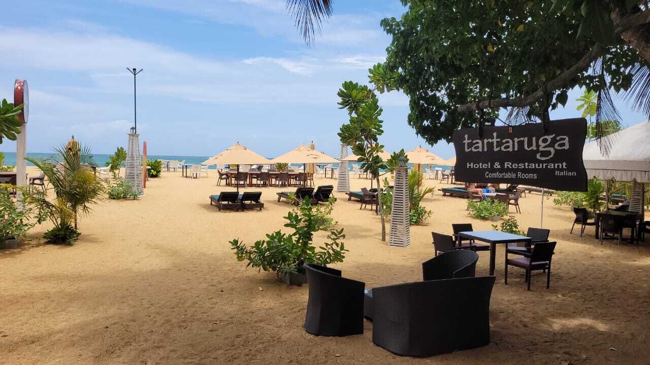 A beachside restaurant with outdoor seating, umbrellas, and a sign indicating it is a 'tartaruga Hotel & Restaurant'.