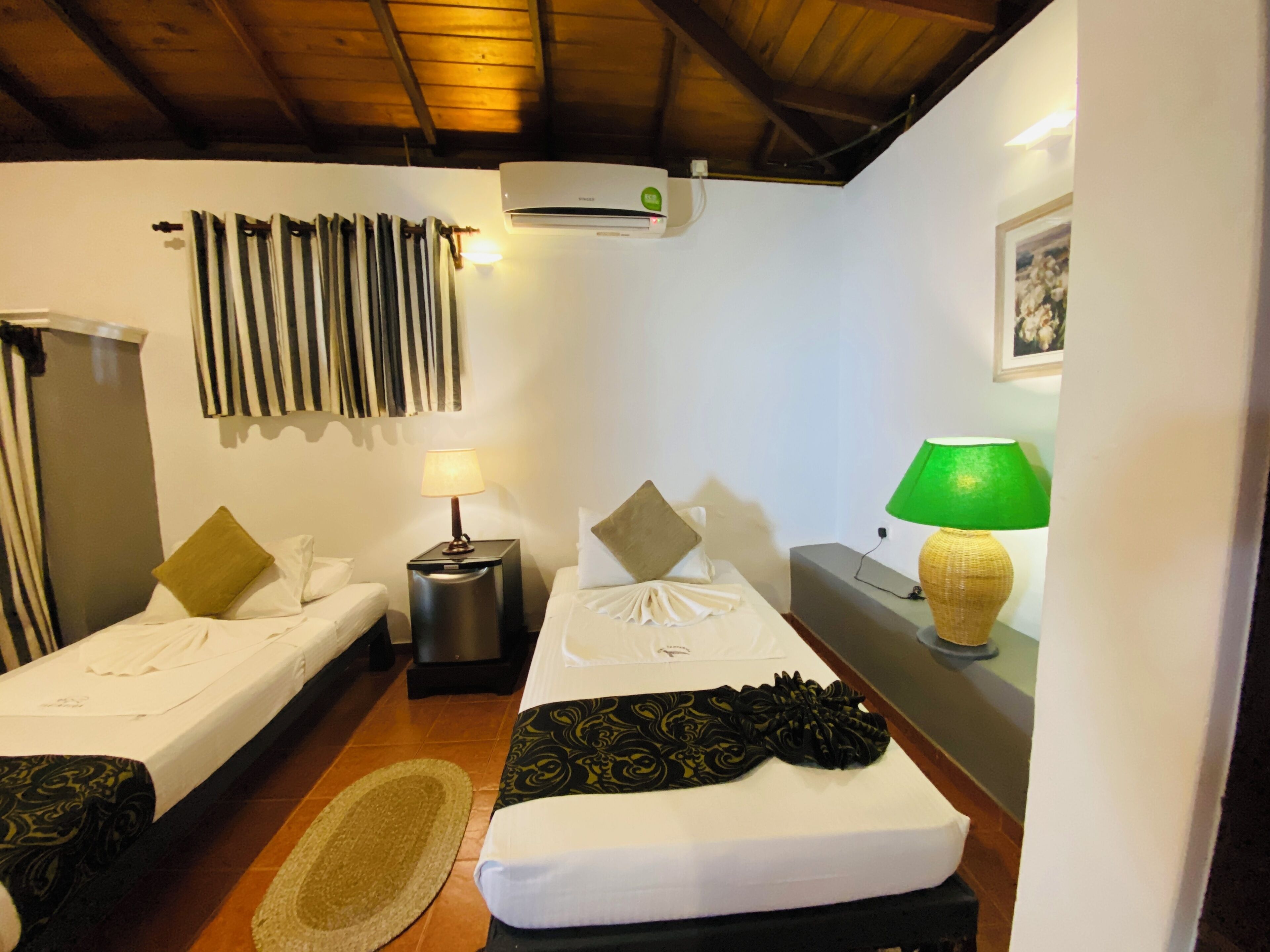 A hotel room with two beds, a small table, a lamp, a wall-mounted air conditioner, and a framed picture on the wall.