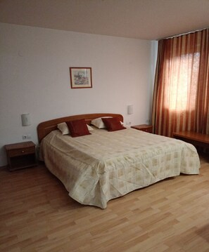 Een slaapkamer met een groot bed, houten vloer, nachtkastje, wandlamp en een raam met gordijnen.