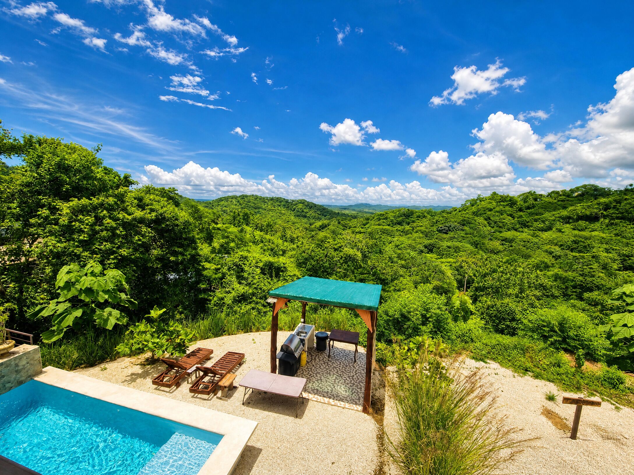 A pool area with lounge chairs and a barbecue grill, surrounded by lush greenery and a clear blue sky.