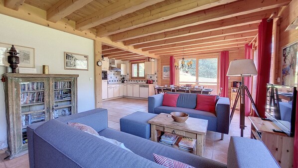L’intérieur d’un chalet accueillant présente un espace de vie avec un canapé bleu, une table basse en bois et une bibliothèque. On aperçoit à l’arrière-plan une cuisine avec des meubles en bois et un coin repas avec une table et des chaises.