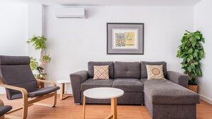 Ein modernes Wohnzimmer mit einem grauen Ecksofa, einem Stuhl mit Holzrahmen, einem kleinen weißen Tisch und zwei Zimmerpflanzen.