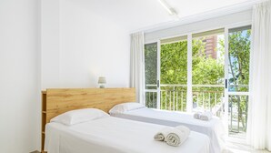 Habitación de hotel con dos camas, cabecero de madera, ventana con vistas a zonas verdes y un edificio, y una pequeña mesa con lámpara.