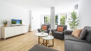 Una sala de estar moderna con un televisor de pantalla plana sobre un mueble blanco, dos sofás grises con cojines naranjas, una mesita blanca con una planta y un ventanal con vistas a zonas verdes.