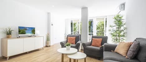 Uma sala de estar moderna com uma TV de tela plana em um armário branco, dois sofás cinzas com almofadas laranja, uma mesa branca pequena com uma planta e uma janela grande com vista para vegetação.