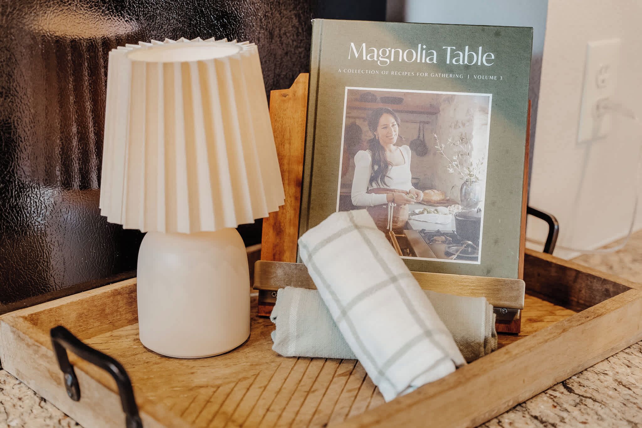 Ein Holztablett mit einer weißen Lampe, einem Buch mit dem Titel 'Magnolia Table' und einem gefalteten weißen Handtuch.