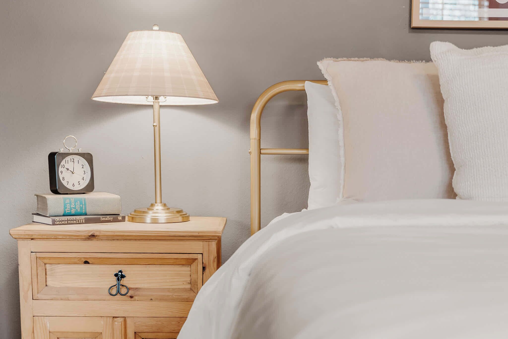 A neatly made bed with white bedding, a bedside lamp, a clock, and a small wooden nightstand with a drawer.