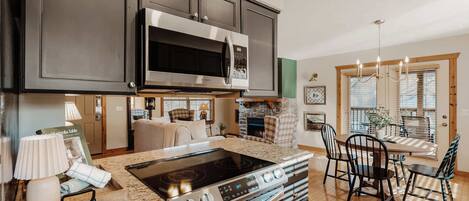 A kitchen with dark cabinets, a stainless steel microwave, a black stove, and a dining area with a table and chairs.