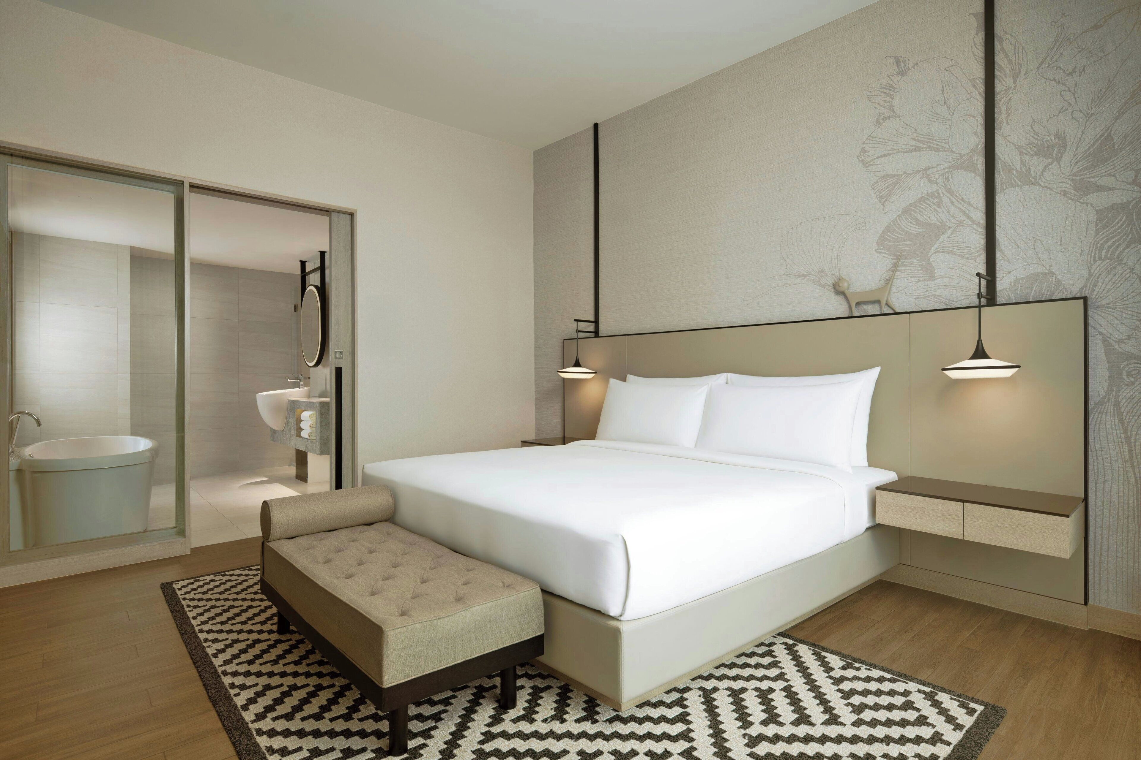 Une chambre moderne avec un grand lit, une table de chevet, une commode, un banc, une cabine de douche en verre et un miroir fixé au mur.