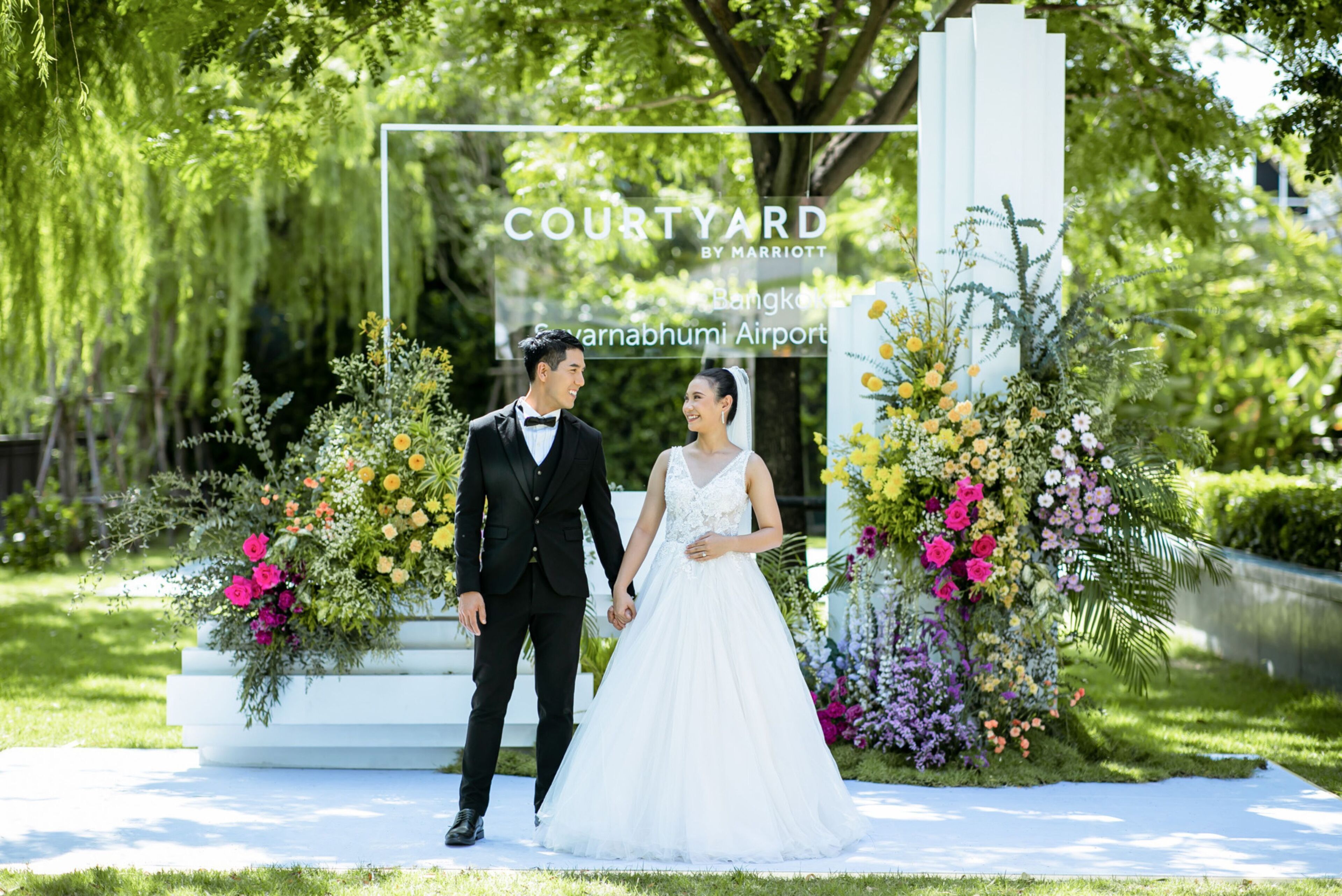 A couple holding hands in front of a floral arch with the text "COURTYARD BY MARRIOTT" and "Bangkok Airport".