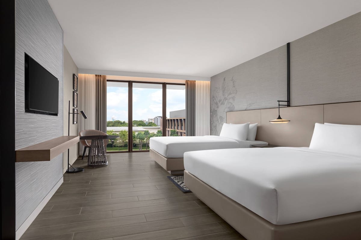 Ett modernt hotellrum med två sängar, en platt-TV som är monterad på väggen, ett litet bord med stolar och ett stort fönster med utsikt över staden.