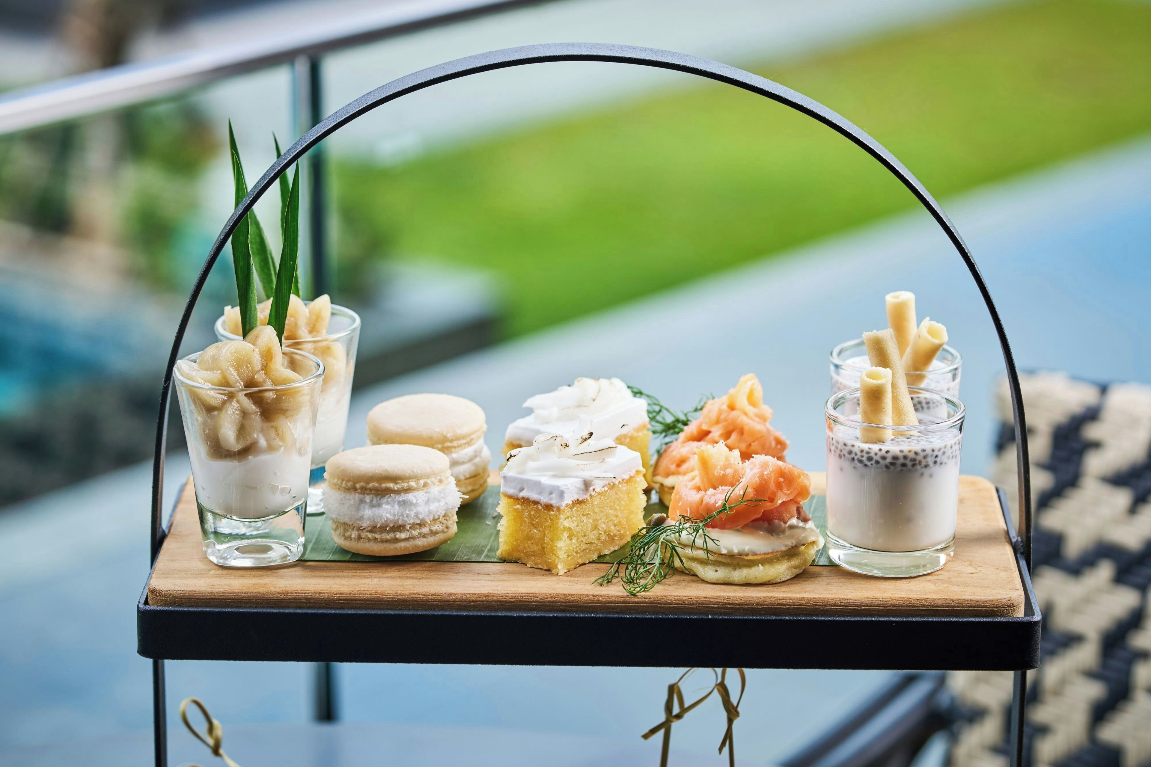 A selection of desserts on a tray, including macarons, a slice of cake, and a glass with a creamy beverage, set against a blurred outdoor background.