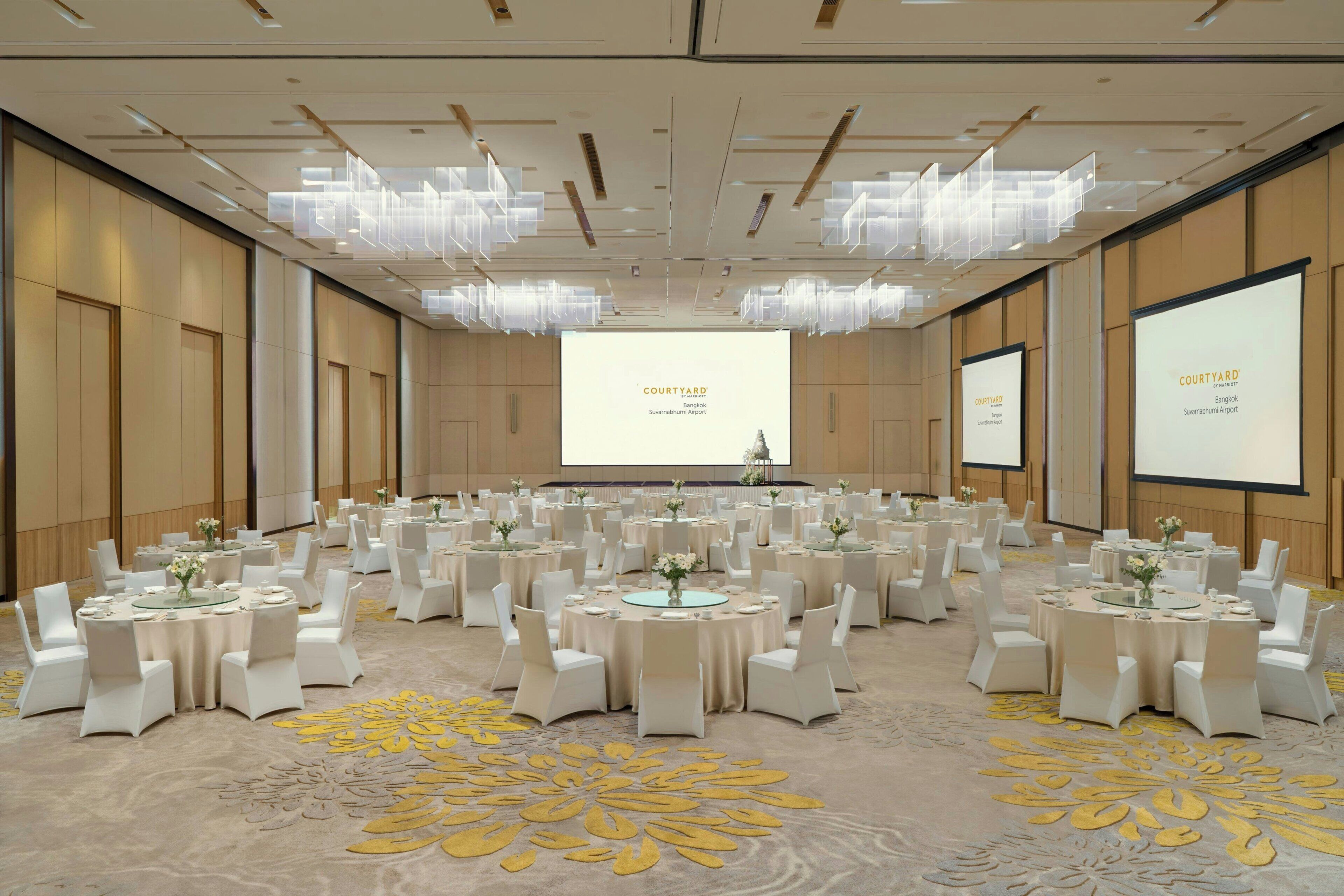A large conference room with multiple round tables, white chairs, and a large screen displaying text.