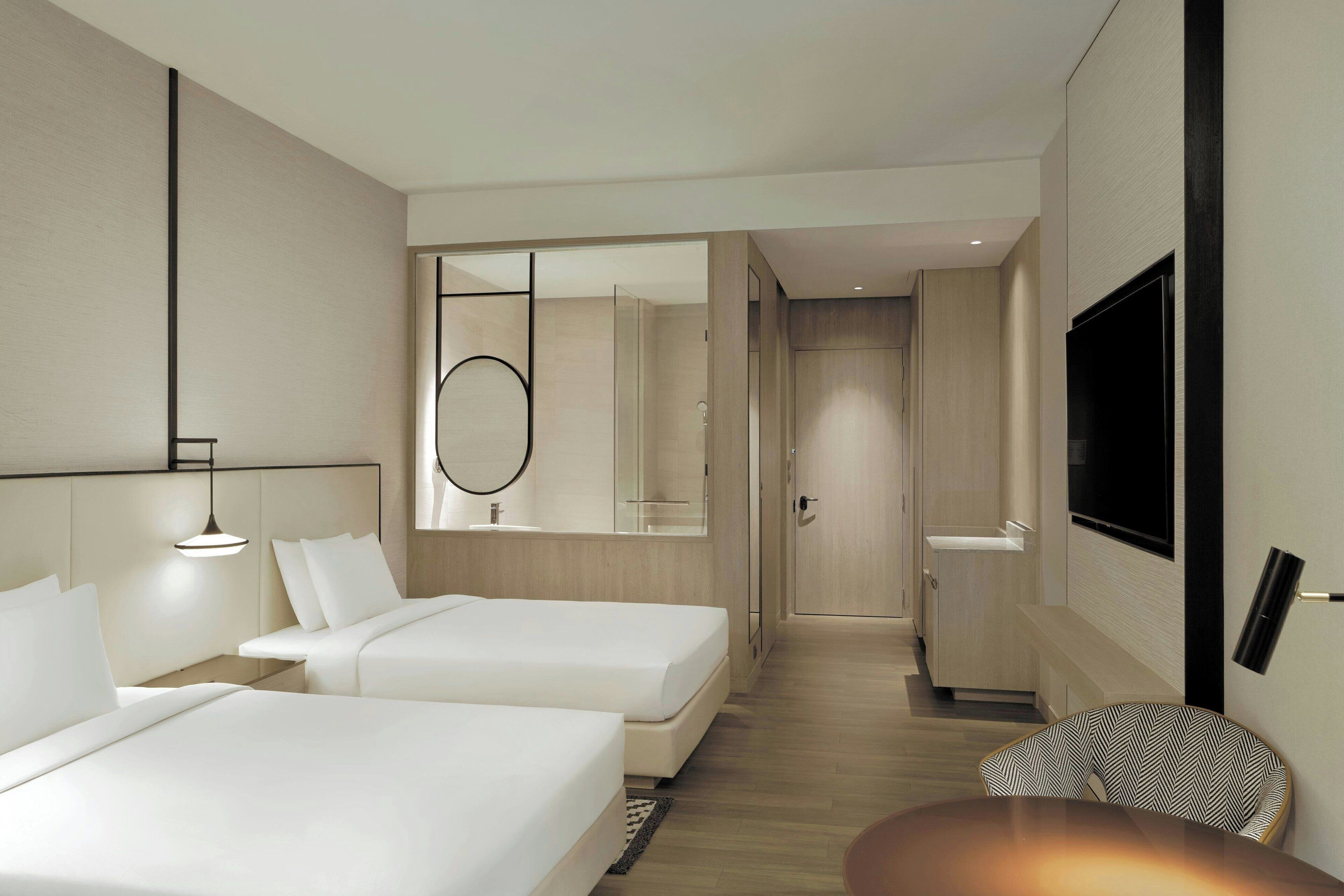 A modern hotel room with two beds, a TV, a mirror, and a small seating area.