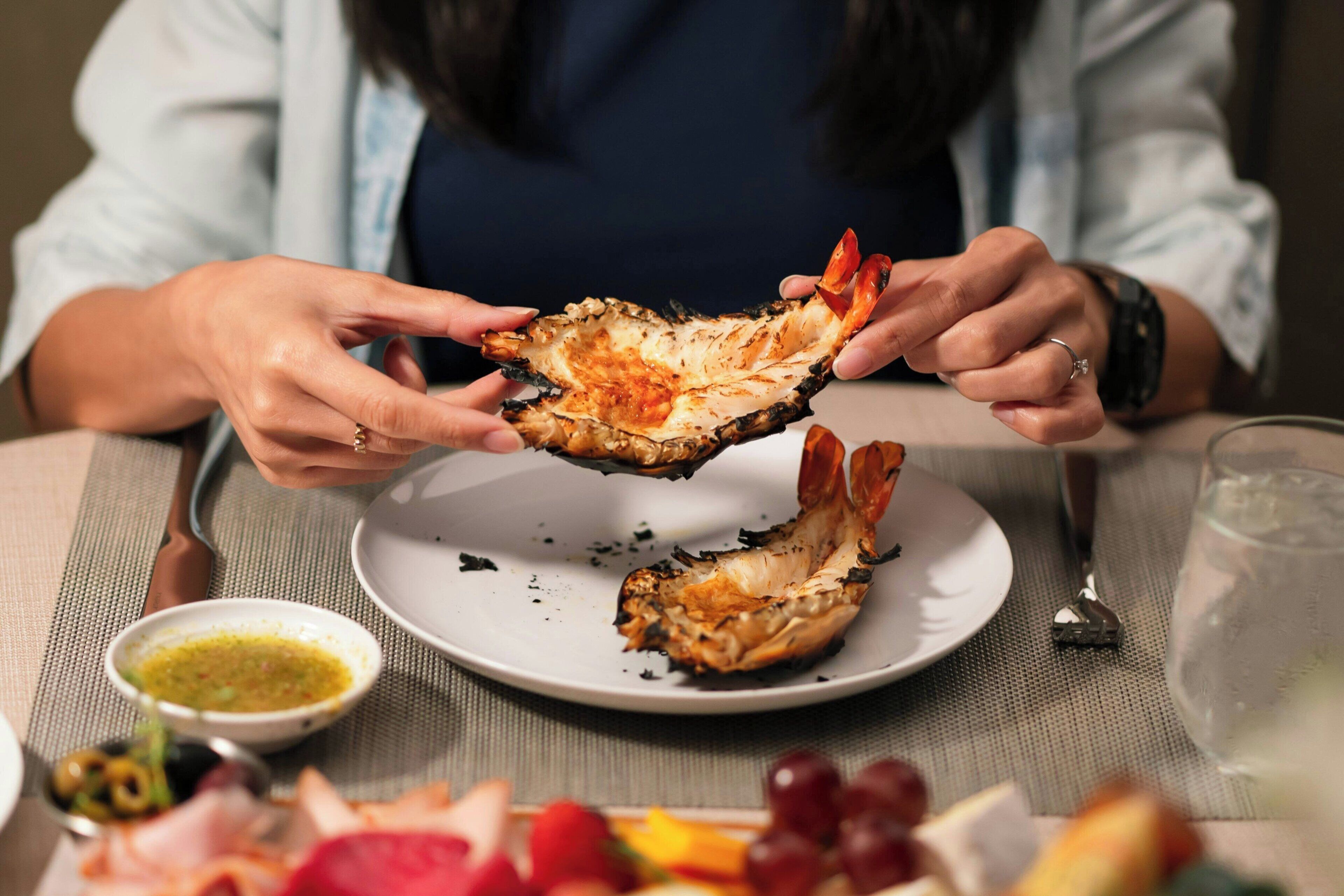 A person holding a cooked lobster shell, a plate with remnants of food, a small bowl of sauce, and a glass of water on a table.
