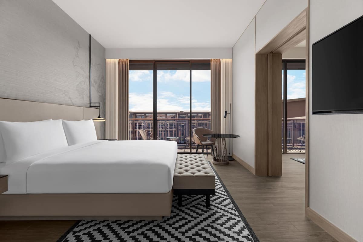 Ett modernt hotellrum med en stor säng, utsikt över staden, en balkong med sittplatser och en platt-TV.