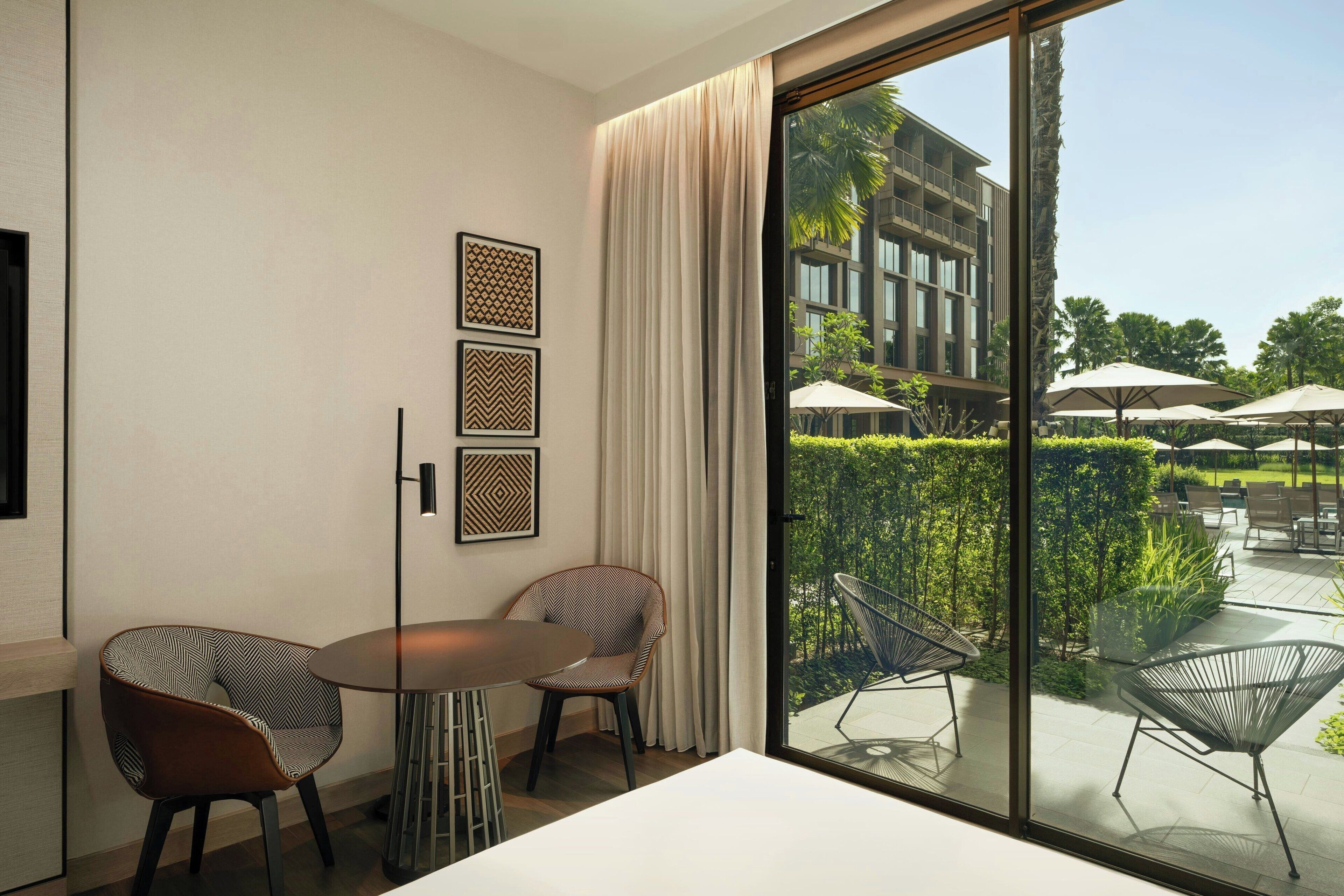 A modern hotel room with a large sliding glass door leading to an outdoor patio area with umbrellas and seating.
