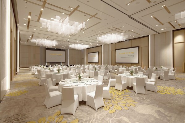 A large, well-lit conference room with multiple round tables and white chairs, featuring a carpeted floor and a modern chandelier.