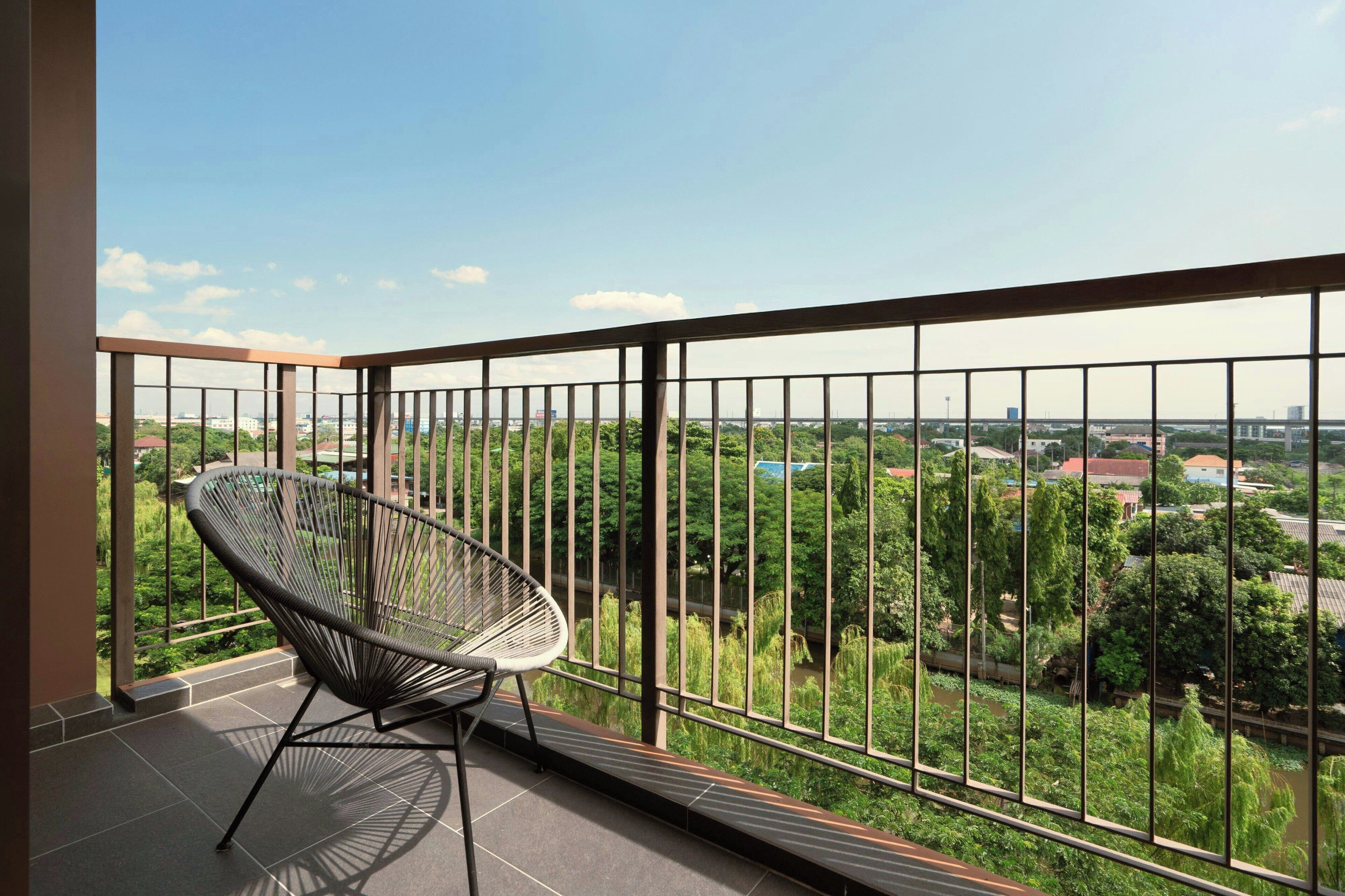 A balcony with a chair, a metal railing, and a view of greenery and buildings.
