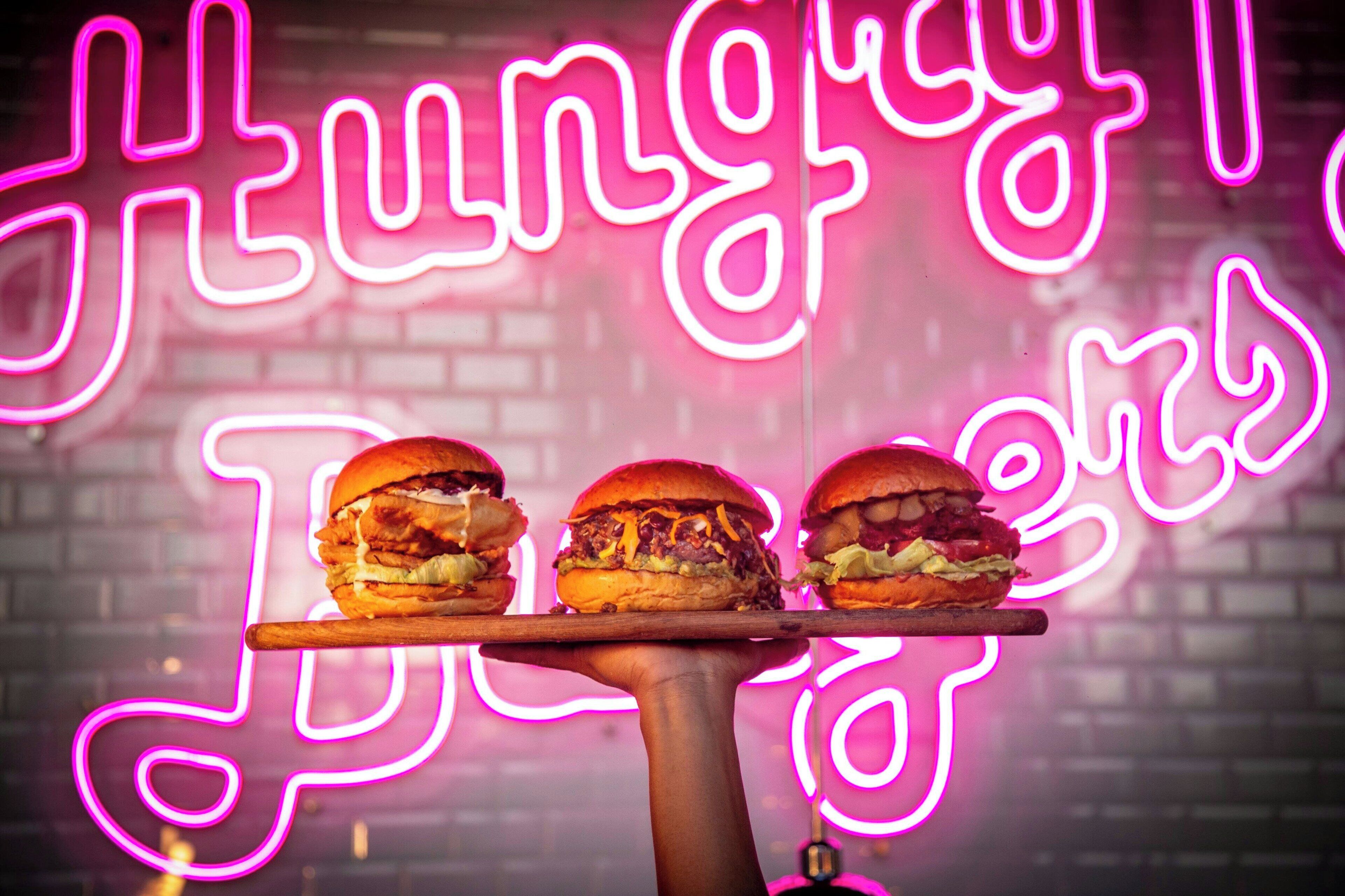 Una mano sostiene una bandeja con tres hamburguesas frente a un letrero de neón.