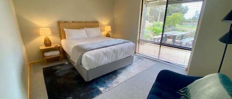 Une chambre à coucher comprenant un lit, des lampes de chevet, une chaise, une grande fenêtre avec des stores et une vue sur la verdure à l’extérieur.