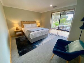 A bedroom with a bed, bedside lamps, a chair, a large window with blinds, and a view of greenery outside.